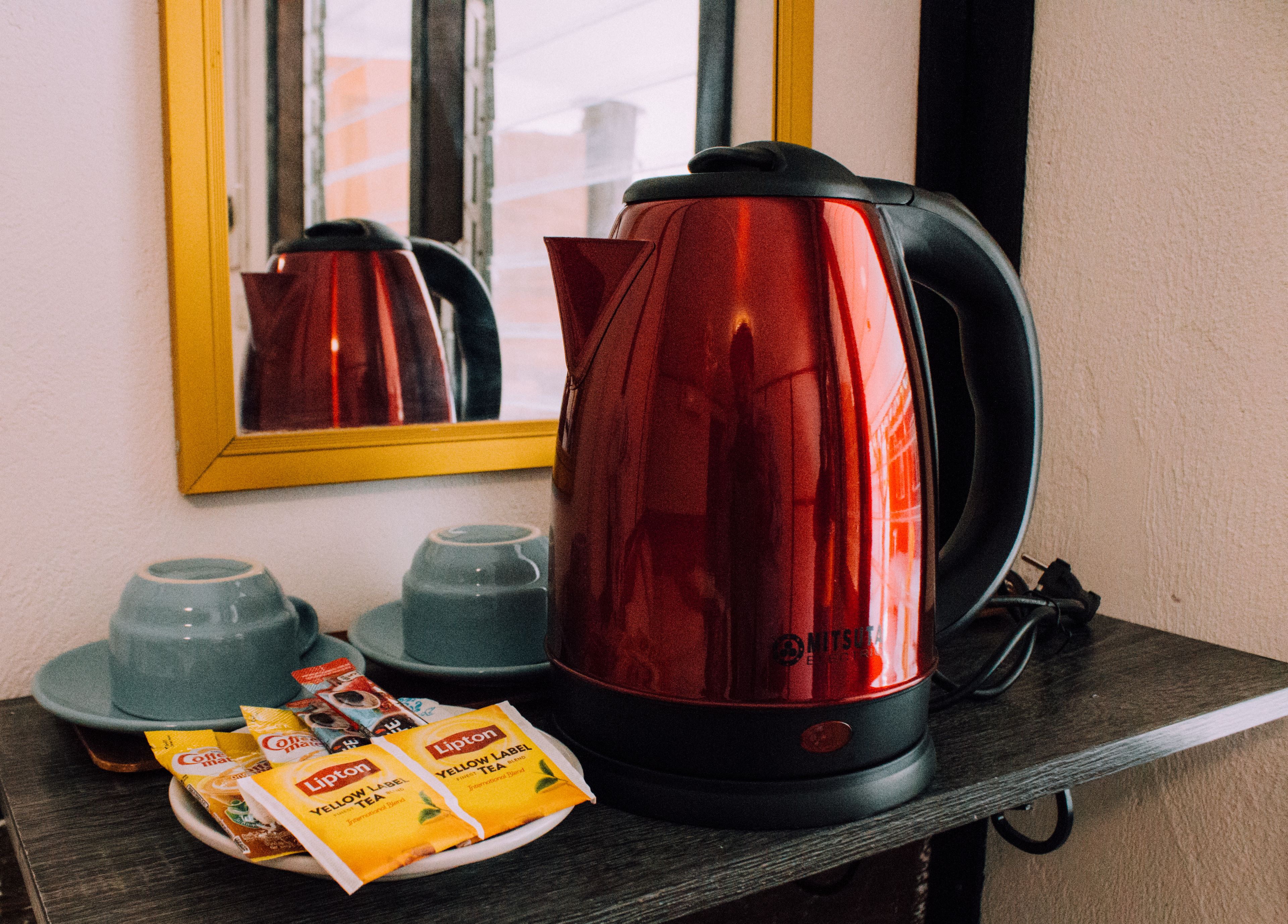 A red electric kettle on a shelf, a plate with Lipton tea packets, and two blue cups.