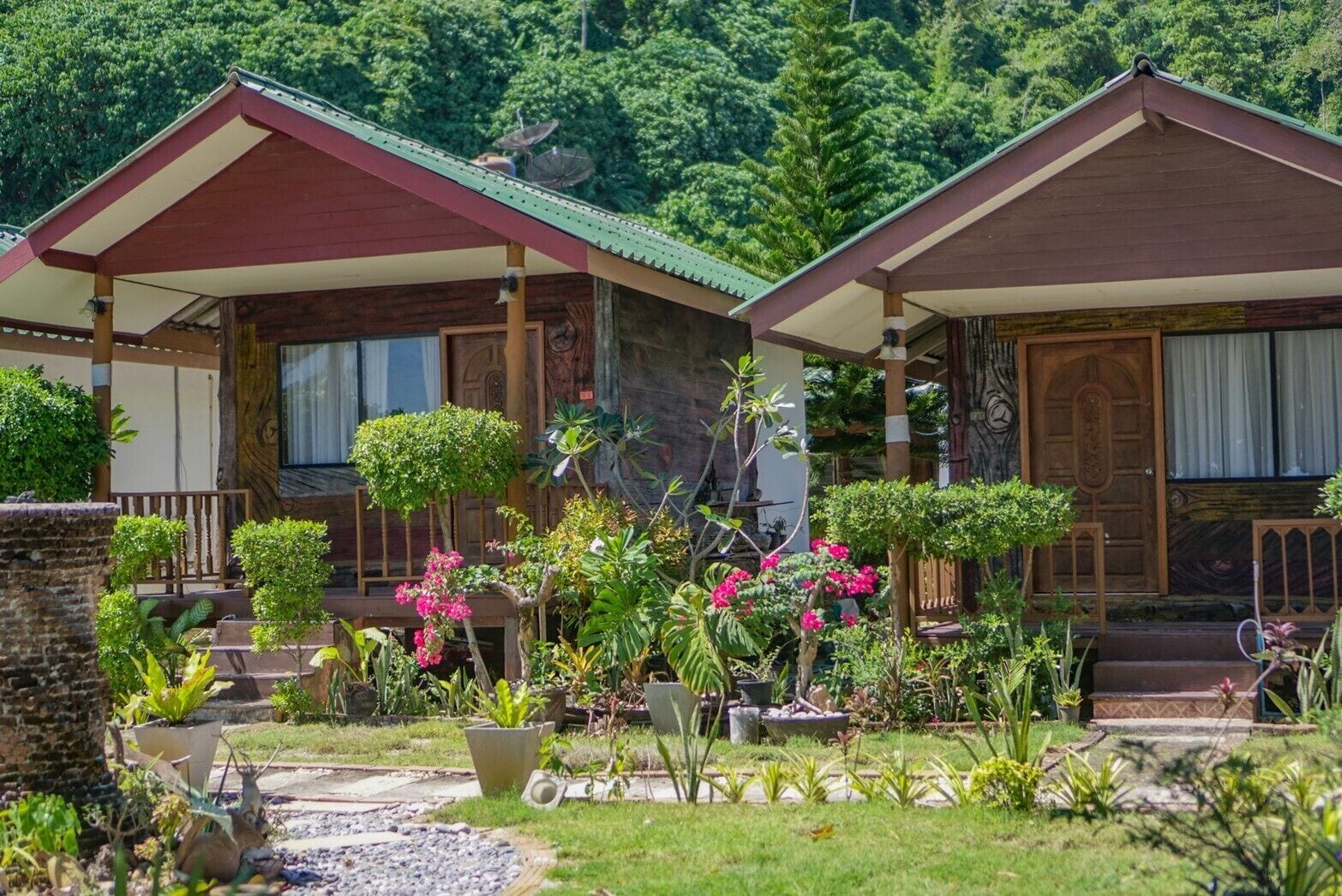 Two wooden cabins with red roofs, surrounded by lush greenery and flowering plants.