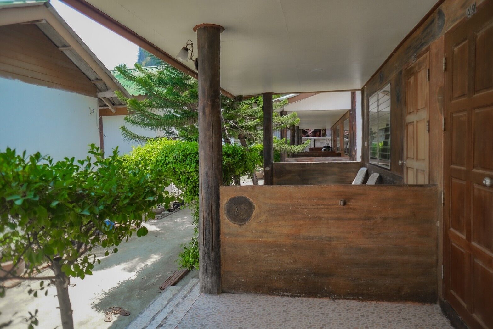 Un espace extérieur abrité, doté d’un banc en bois, d’un sol carrelé et offrant une vue sur un bâtiment avec une porte de garage.