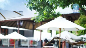 A poolside area with lounge chairs and umbrellas, a pool, and a building with a thatched roof in the background. - Ozz Hotel - Kuta Bali (Kuta)
