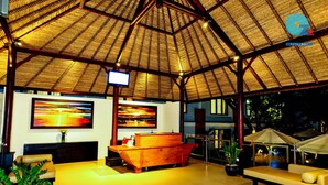 A spacious lounge area with a thatched roof, comfortable seating, a flat-screen TV, and artwork on the walls. - Ozz Hotel - Kuta Bali (Kuta)