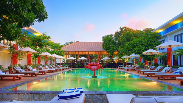 A pool area with lounge chairs and umbrellas, surrounded by trees and a building with a tiled roof. - Ozz Hotel - Kuta Bali (Kuta)