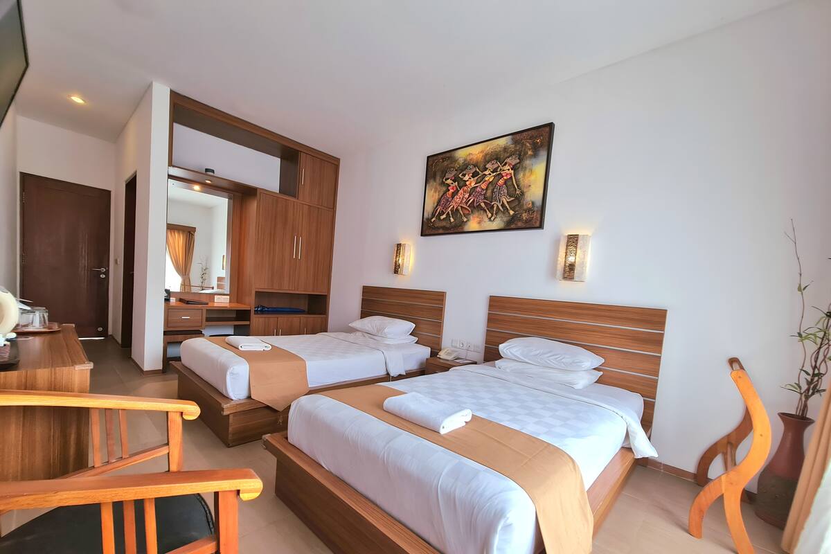 Habitación de hotel con dos camas, cabeceros de madera, un escritorio con una silla, un espejo y un cuadro en la pared.
