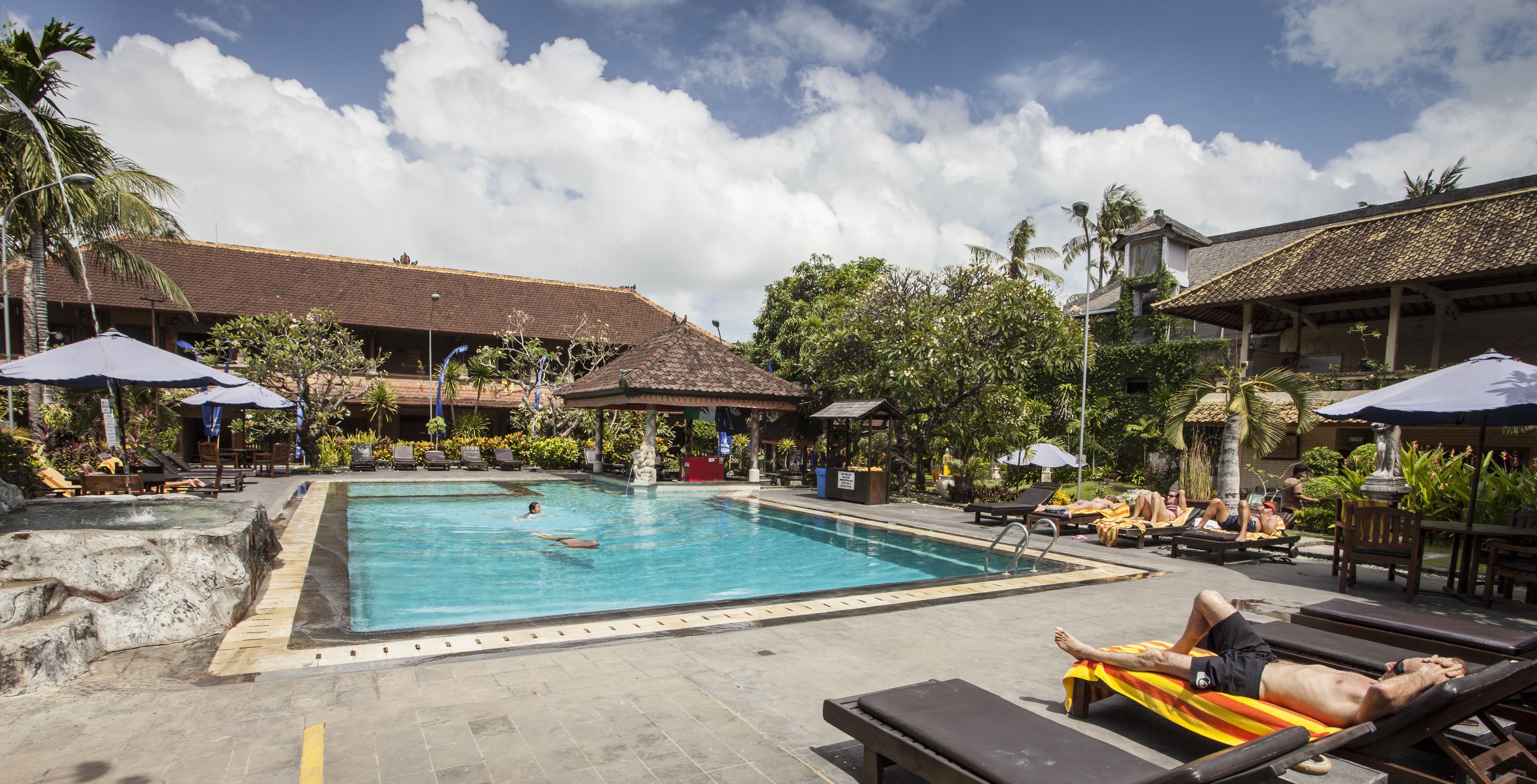 A swimming pool with lounge chairs and umbrellas, surrounded by tropical plants and buildings with tiled roofs.