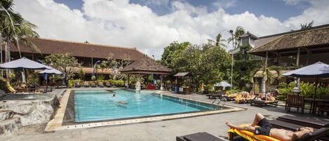 A swimming pool with lounge chairs and umbrellas, surrounded by tropical plants and buildings with tiled roofs.