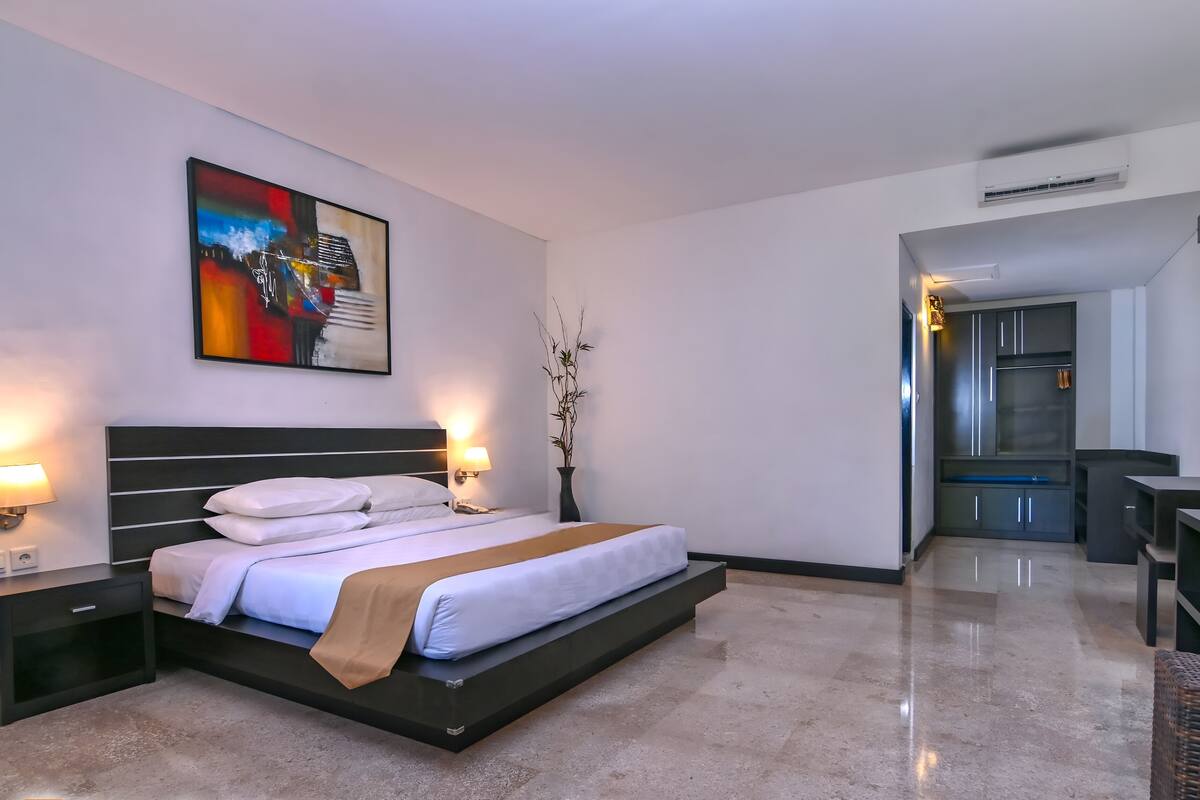 Una habitación de hotel moderna con una cama grande, lámparas de noche, un cuadro y una planta pequeña en maceta.