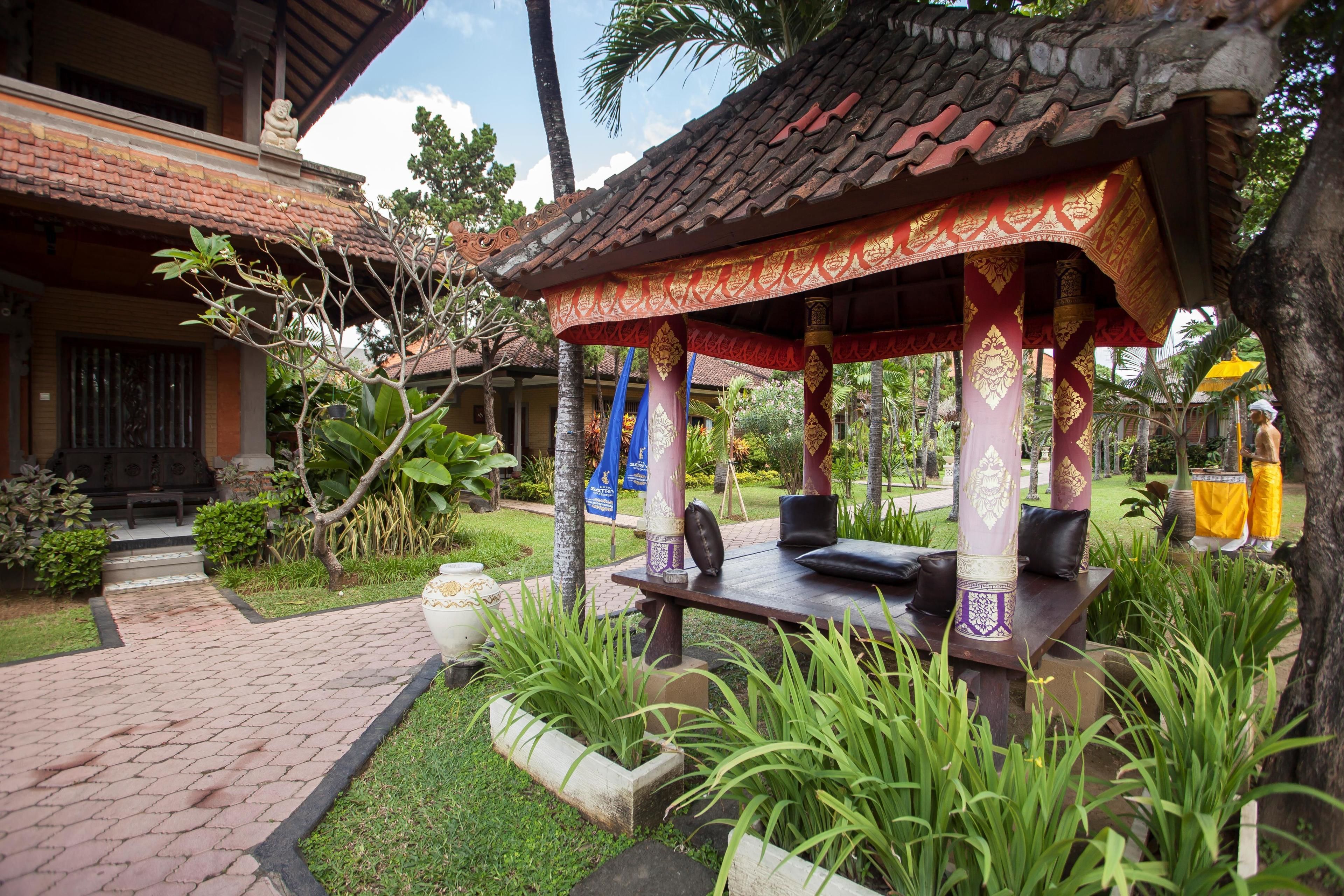 A traditional pavilion with ornate columns and a tiled roof, surrounded by lush greenery and a paved pathway.