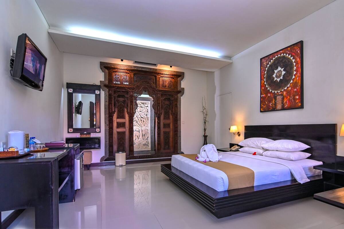 Habitación de hotel con cama, mesitas de noche, escritorio, espejo, puerta de madera decorativa y una obra de arte colgada en la pared.