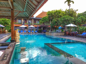 A swimming pool with lounge chairs and umbrellas, surrounded by tropical plants and traditional architecture. - Satriya Cottages (Kuta)