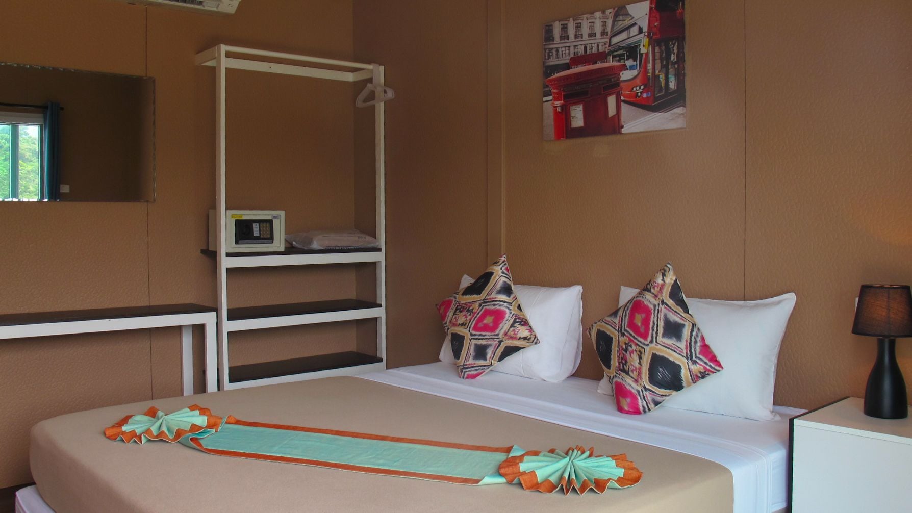 Habitación de hotel con cama, mesita de noche, estante con un reloj y un cuadro colgado en la pared de un autobús.