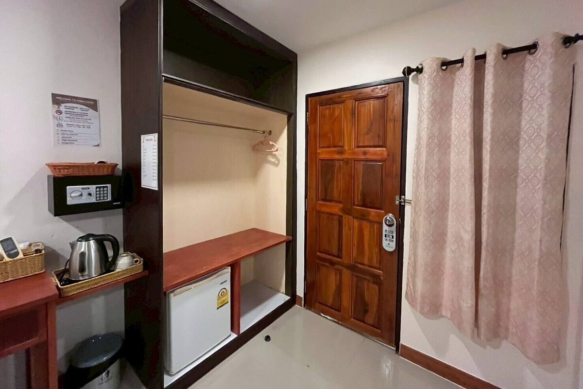 A compact room with a wooden door, a small refrigerator, a kettle, a basket, and a shelf with a control panel.