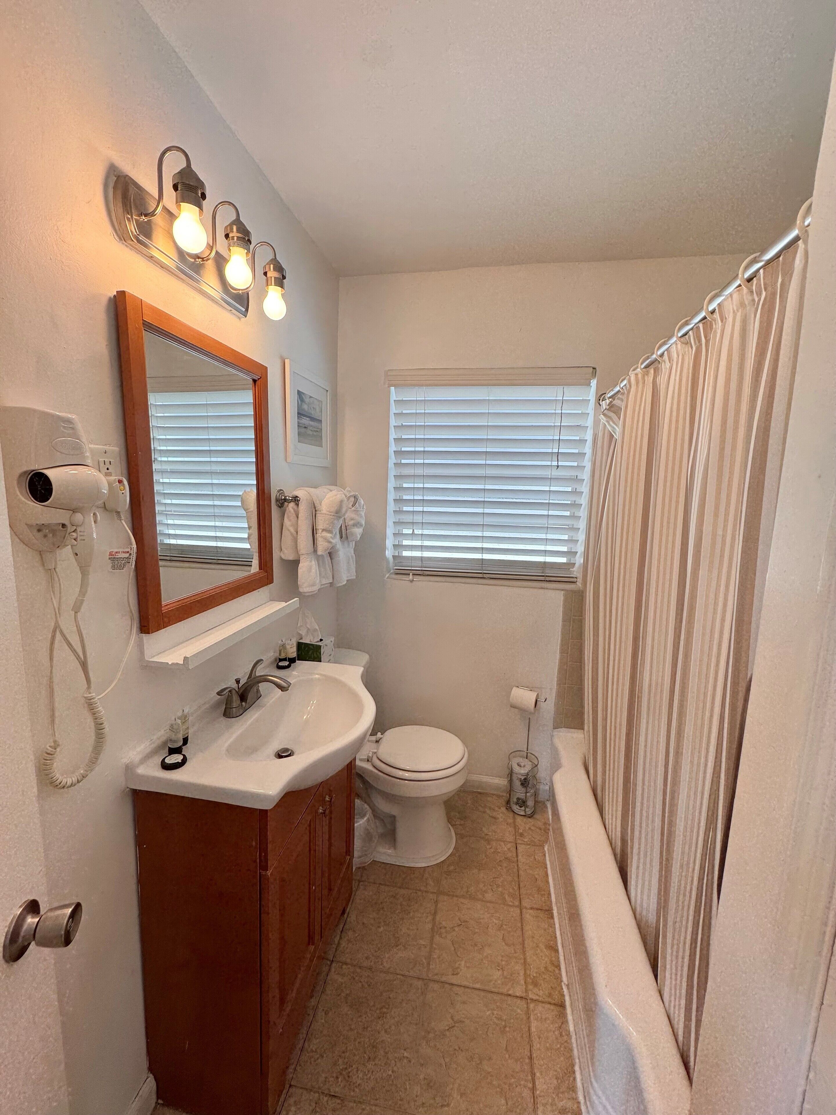 Un baño con lavabo, espejo, secador de pelo, inodoro y ventana con persianas.