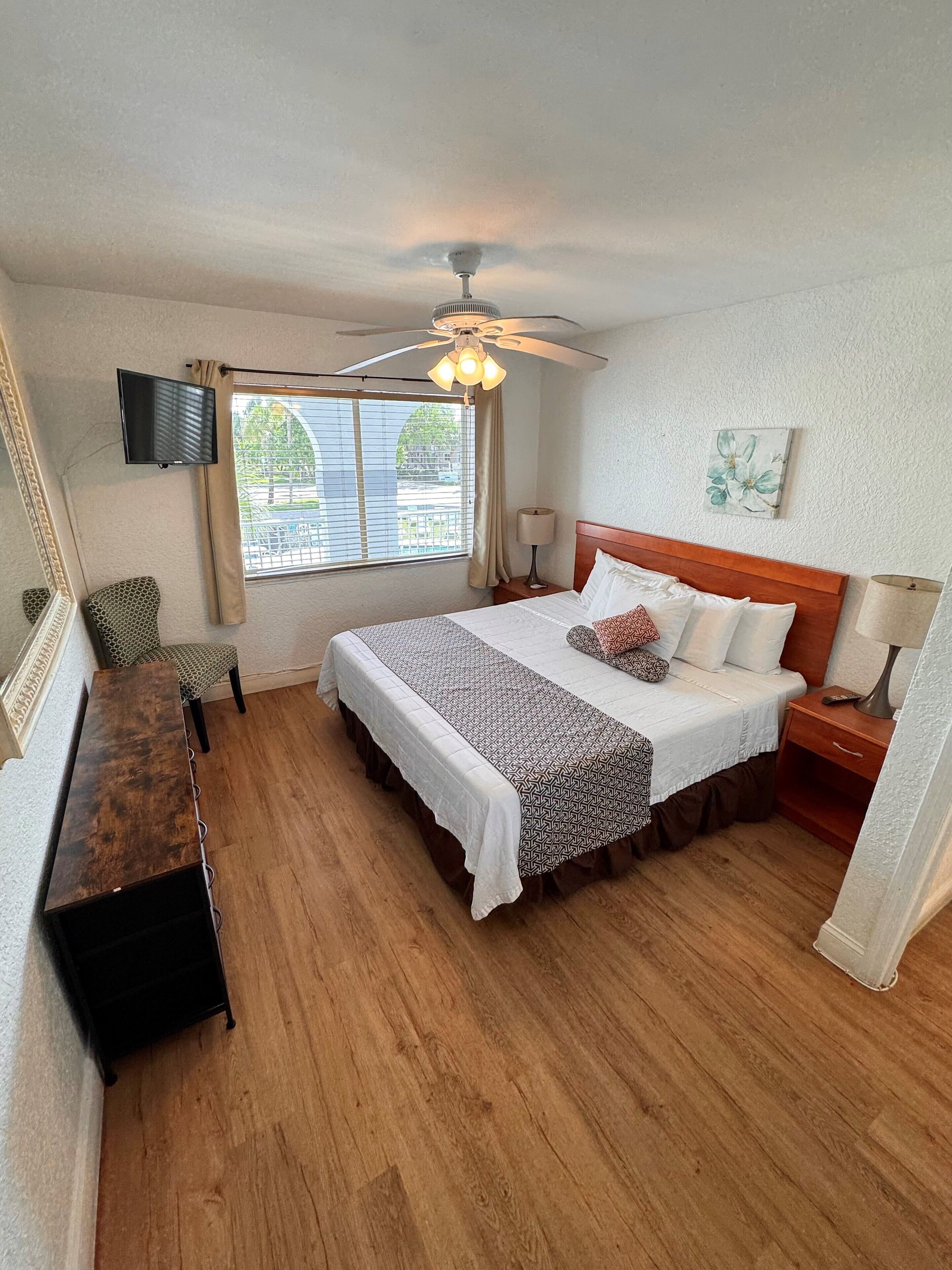 A bedroom with a bed, bedside table, chair, TV, ceiling fan, window, and artwork.