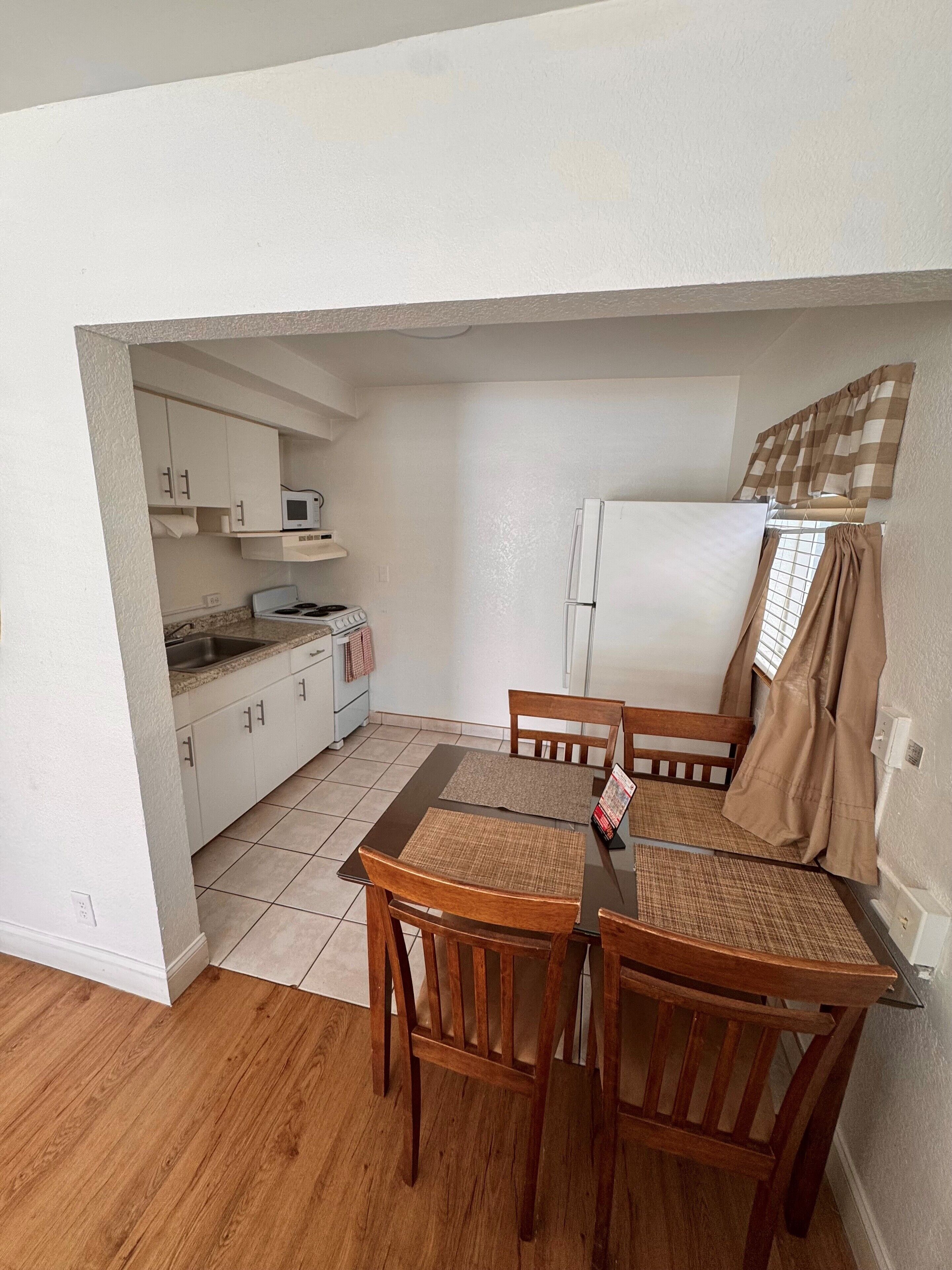 A compact kitchen with white cabinets, a microwave, a stove, and a refrigerator. There is a dining table with two chairs and a placemat. The floor is tiled, and the walls are plain.