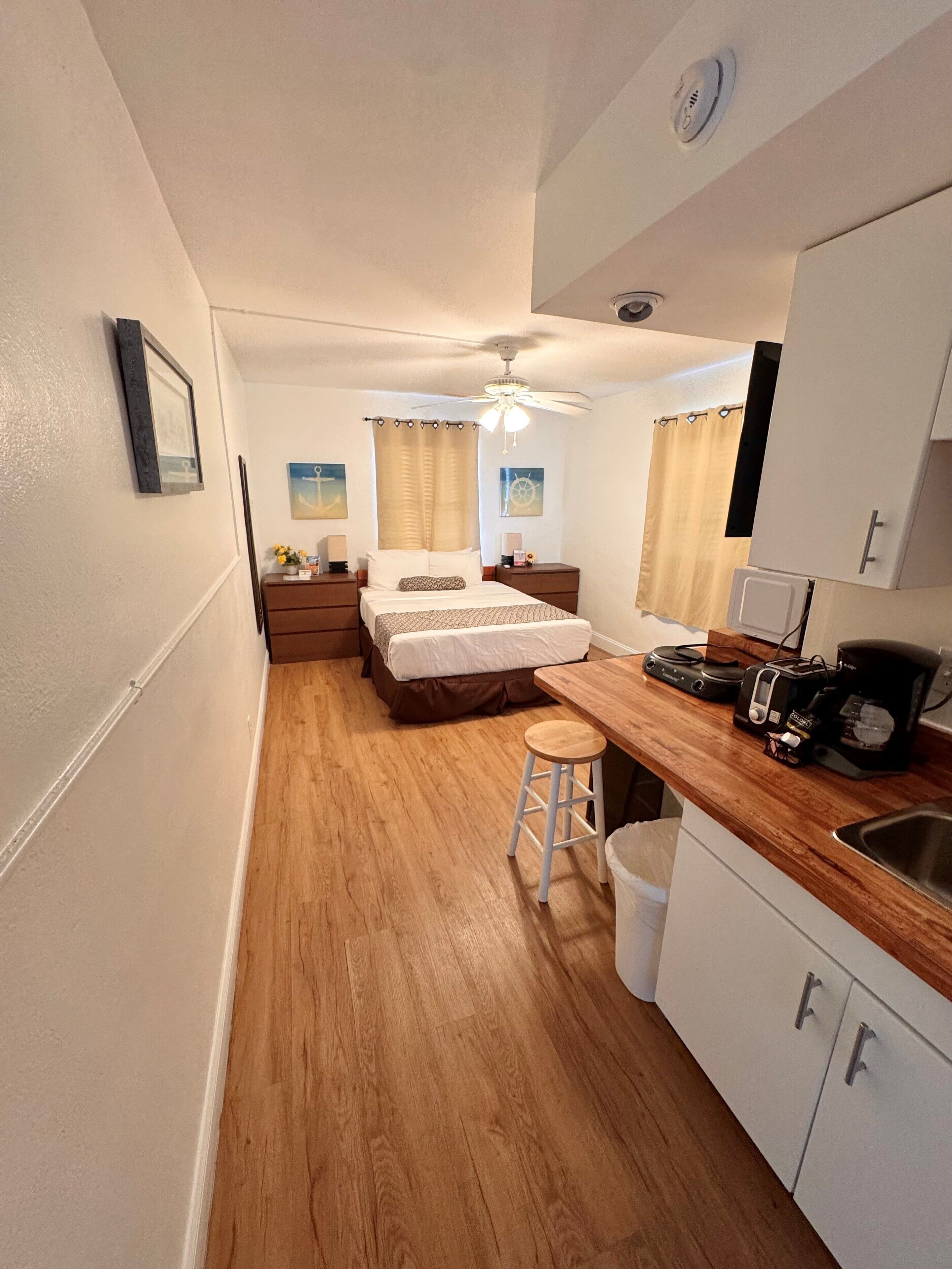 Habitación de hotel compacta con cama, una kitchenette pequeña, ventilador de techo y un taburete.