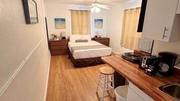 Habitación de hotel compacta con cama, una kitchenette pequeña, ventilador de techo y un taburete.