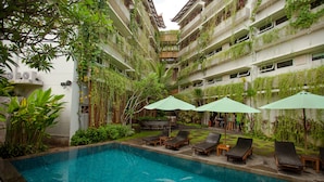 Una piscina con sillones y sombrillas, rodeada de abundante vegetación y un edificio de varios pisos con balcones.