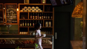Interior de restaurante con una mujer caminando, estantes llenos de botellas y una pizarra anunciando una promoción de balde de cerveza.