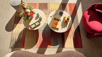 Vista cenital de una habitación con un tapete estampado, una silla roja, una mesa con un libro titulado 'BALI' y una pequeña planta en maceta.