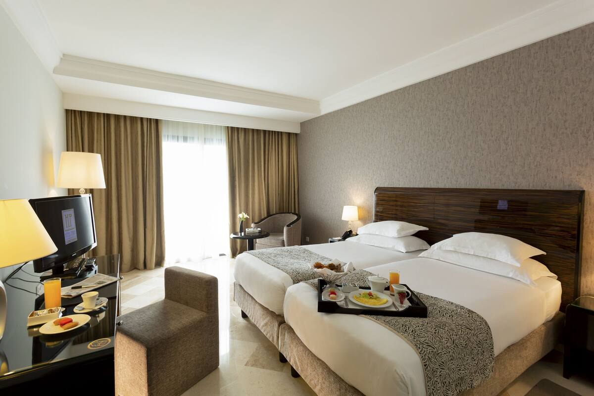 Quarto de hotel com uma cama grande, uma escrivaninha com computador, uma cadeira e uma bandeja com itens de café da manhã.