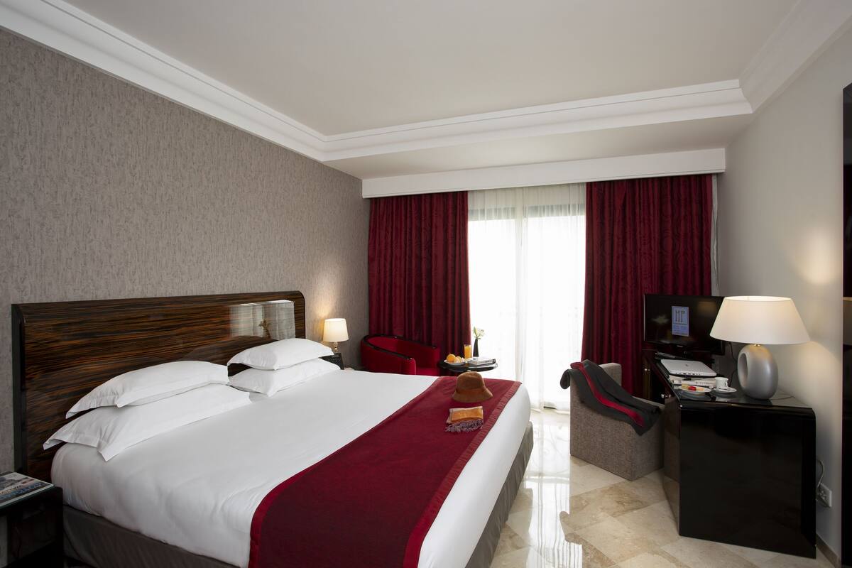 Quarto de hotel com uma cama grande, uma escrivaninha com abajur, uma televisão e uma cortina vermelha.