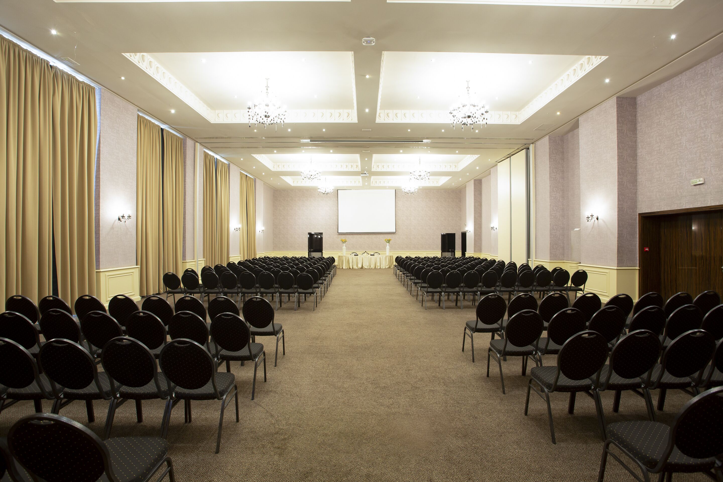 A conference room with rows of chairs facing a presentation screen, chandeliers, and a carpeted floor.