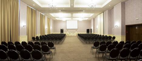 Un salón de conferencias con filas de sillas mirando una pantalla de presentación, candelabros y suelo alfombrado.