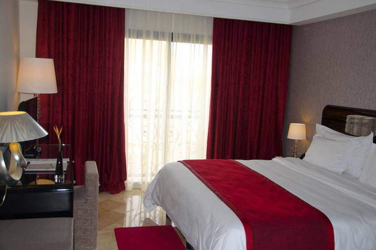 Quarto de hotel com uma cama grande, cortinas vermelhas, uma escrivaninha com abajur e vista pela janela.