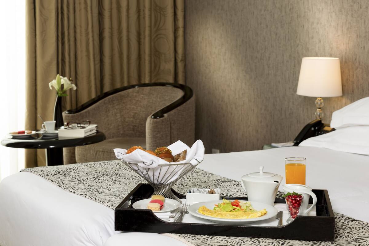 Uma bandeja de café da manhã bem organizada sobre a cama, com um edredom estampado, uma mesa de cabeceira com abajur e uma cadeira com um vaso de flores.
