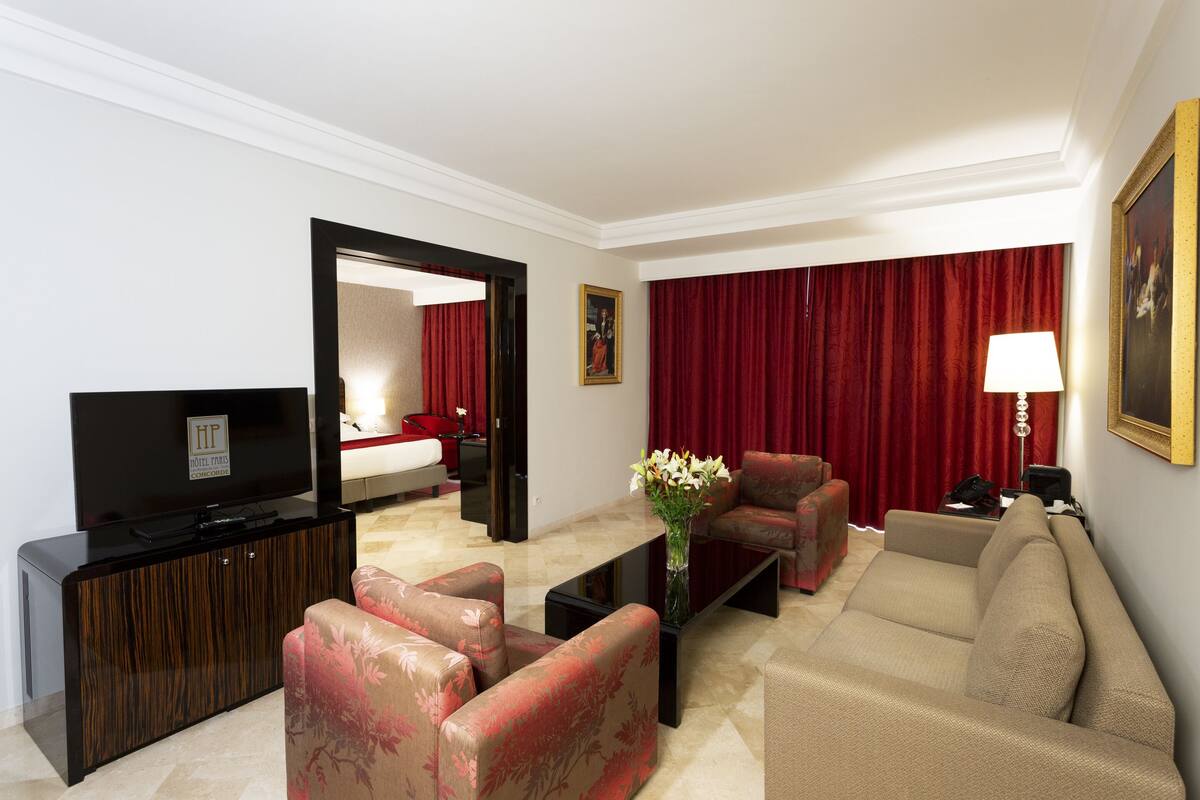 Quarto de hotel com televisão de tela plana, sofá, chaise longue, mesa de centro com um vaso de flores, cortina vermelha e um quadro na parede.