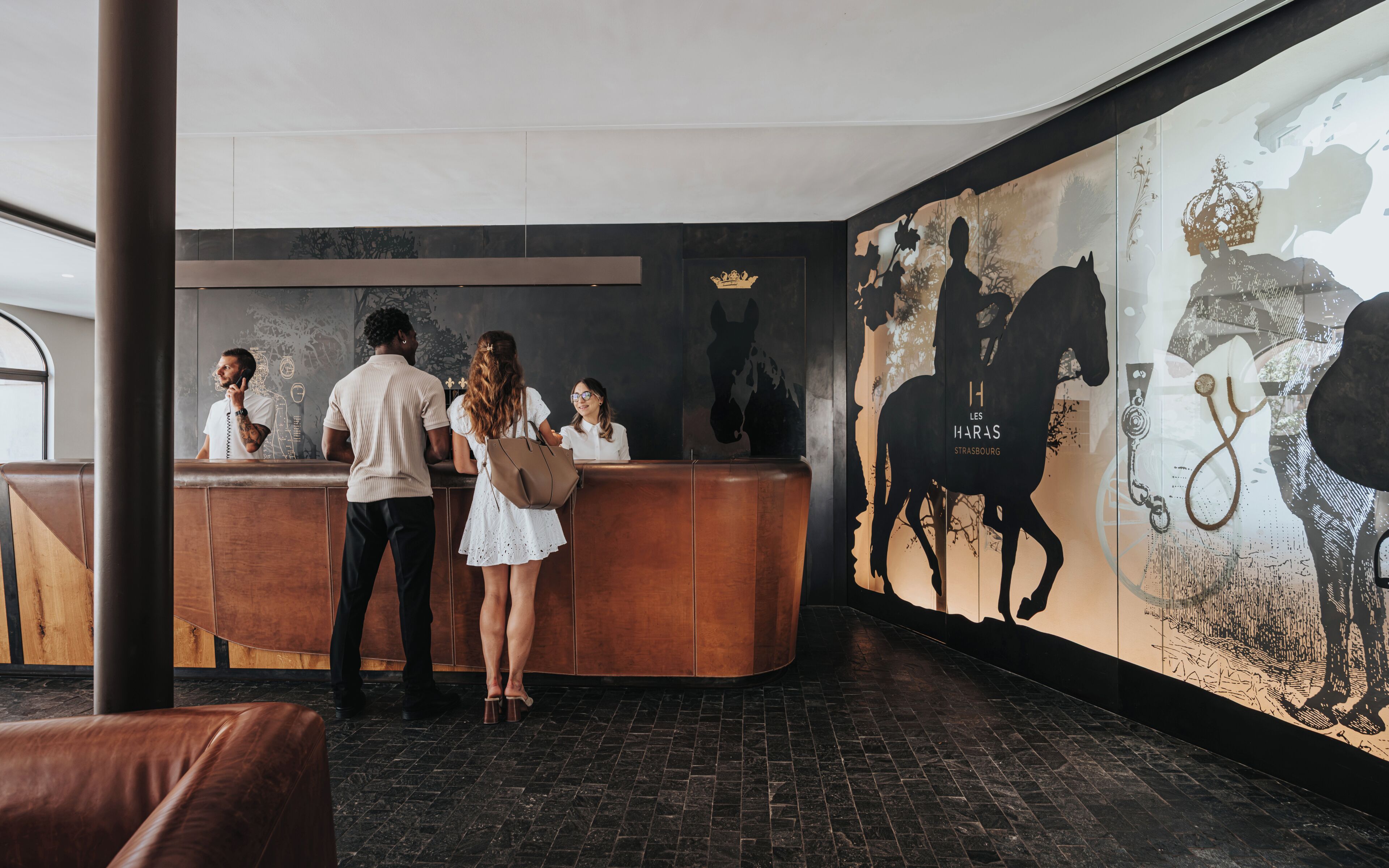 Un espace d’accueil doté d’un comptoir en bois, d’une grande fresque murale représentant des silhouettes de chevaux et d’un sol carrelé de carreaux noirs.