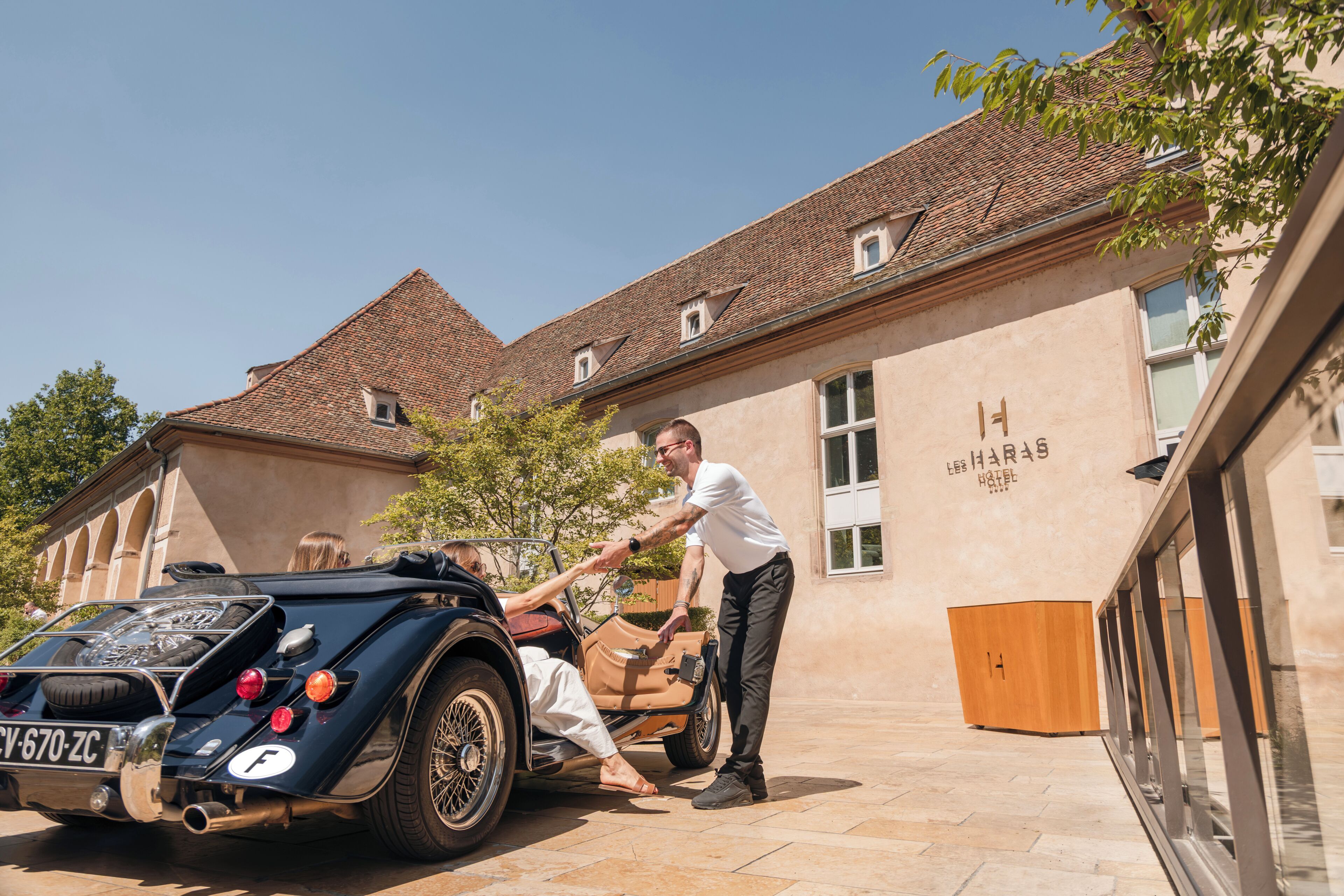 A vintage car parked in front of a building with a tiled roof and a sign indicating a hotel.