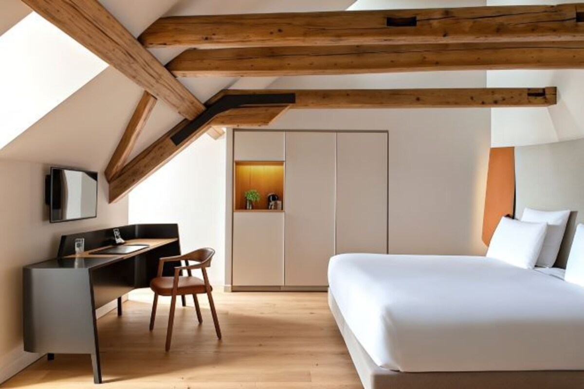 A modern bedroom with a bed, a desk, and a chair. There is a wardrobe and a small plant visible.