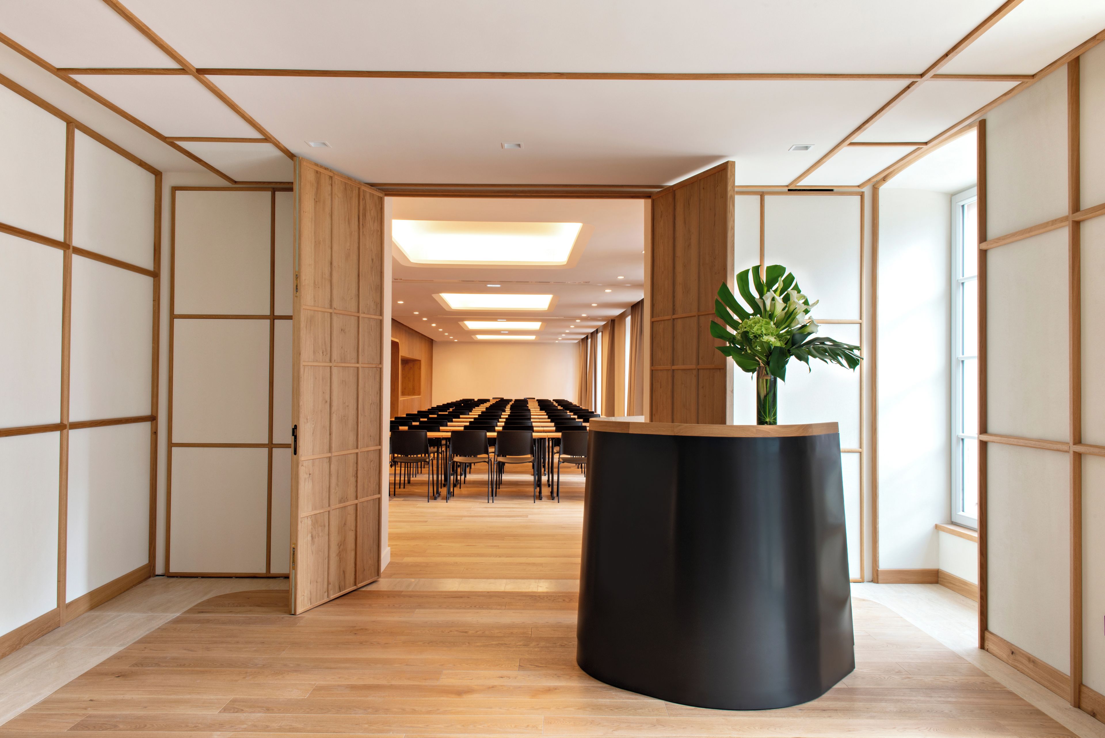 Ein modernes Interieur mit Holzboden, einem schwarzen, zylindrischen Podest und einer Reihe von Stühlen im Hintergrund.