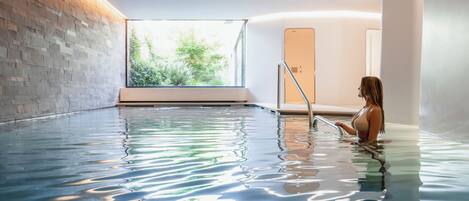 A large indoor swimming pool with clear water, a wooden wall, and a view of greenery outside.