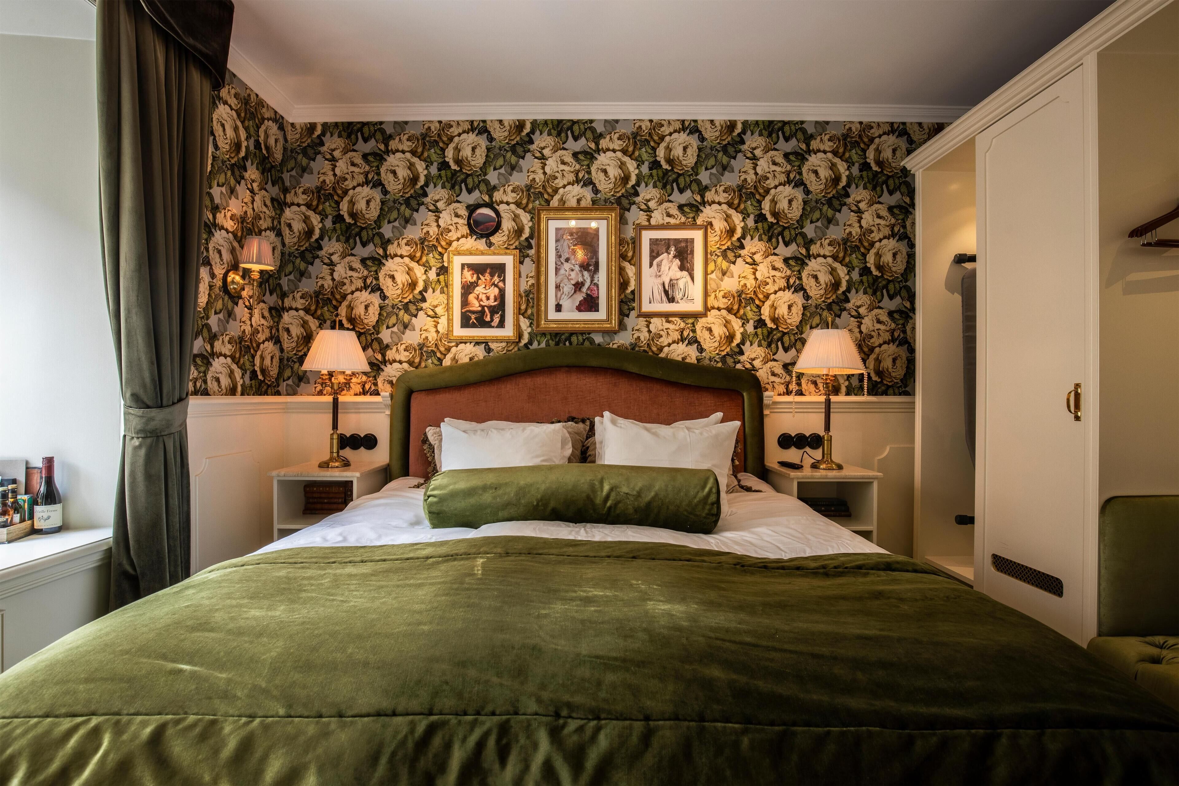 Una cama bien hecha con una colcha verde, flanqueada por dos mesitas de noche con lámparas, un papel tapiz floral y cuadros enmarcados en la pared.