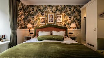 Una cama bien hecha con una colcha verde, flanqueada por dos mesitas de noche con lámparas, un papel tapiz floral y cuadros enmarcados en la pared.