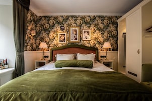 Una cama bien hecha con una colcha verde, flanqueada por dos mesitas de noche con lámparas, un papel tapiz floral y cuadros enmarcados en la pared.