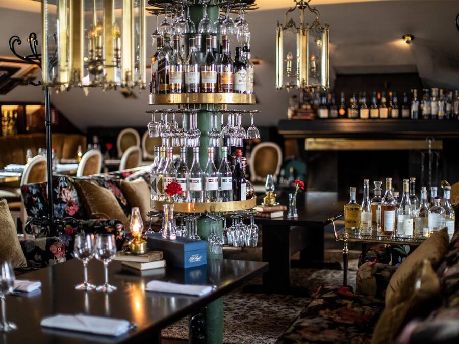 A bar area with a wine rack, glasses, and bottles. There are tables with chairs, a chandelier, and a fireplace in the background.