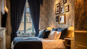 Habitación de hotel con una cama grande, cortinas oscuras, ventana con vistas a edificios y una pared decorada con cuadros.