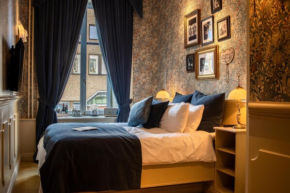 A hotel room with a large bed, dark curtains, a window with a view of buildings, and a wall decorated with framed pictures. - Hotel Pigalle (Gothenburg)