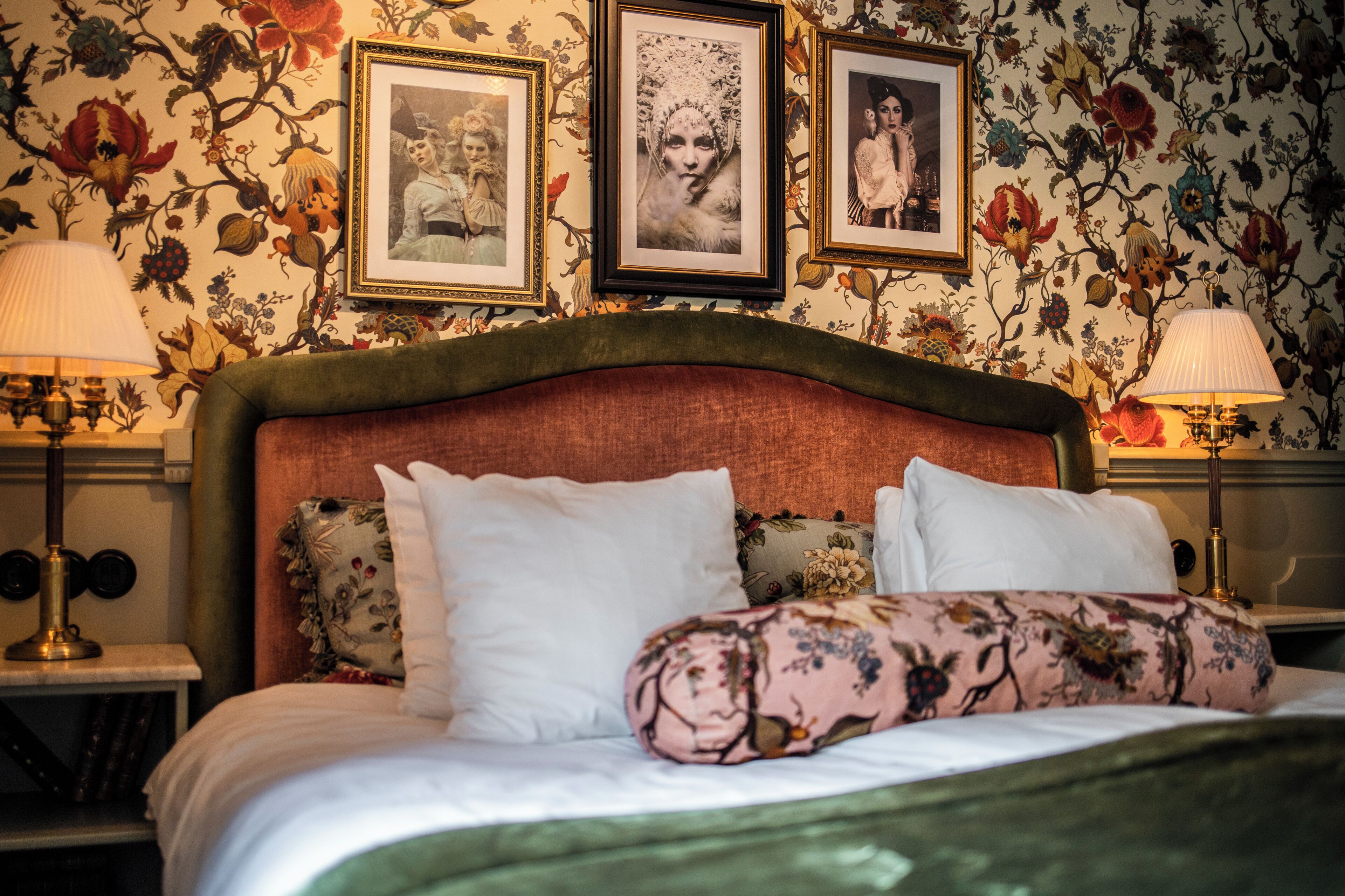 Una cama con almohadas estampadas con motivos florales, un cabecero tapizado en verde y cuadros enmarcados en la pared.