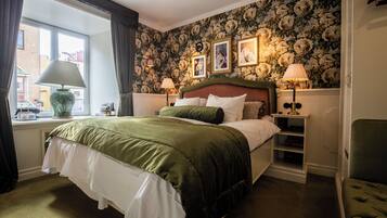Quarto com cama grande, duas luminárias de cabeceira, mesa de cabeceira, uma cadeira, janela com cortinas e papel de parede floral.