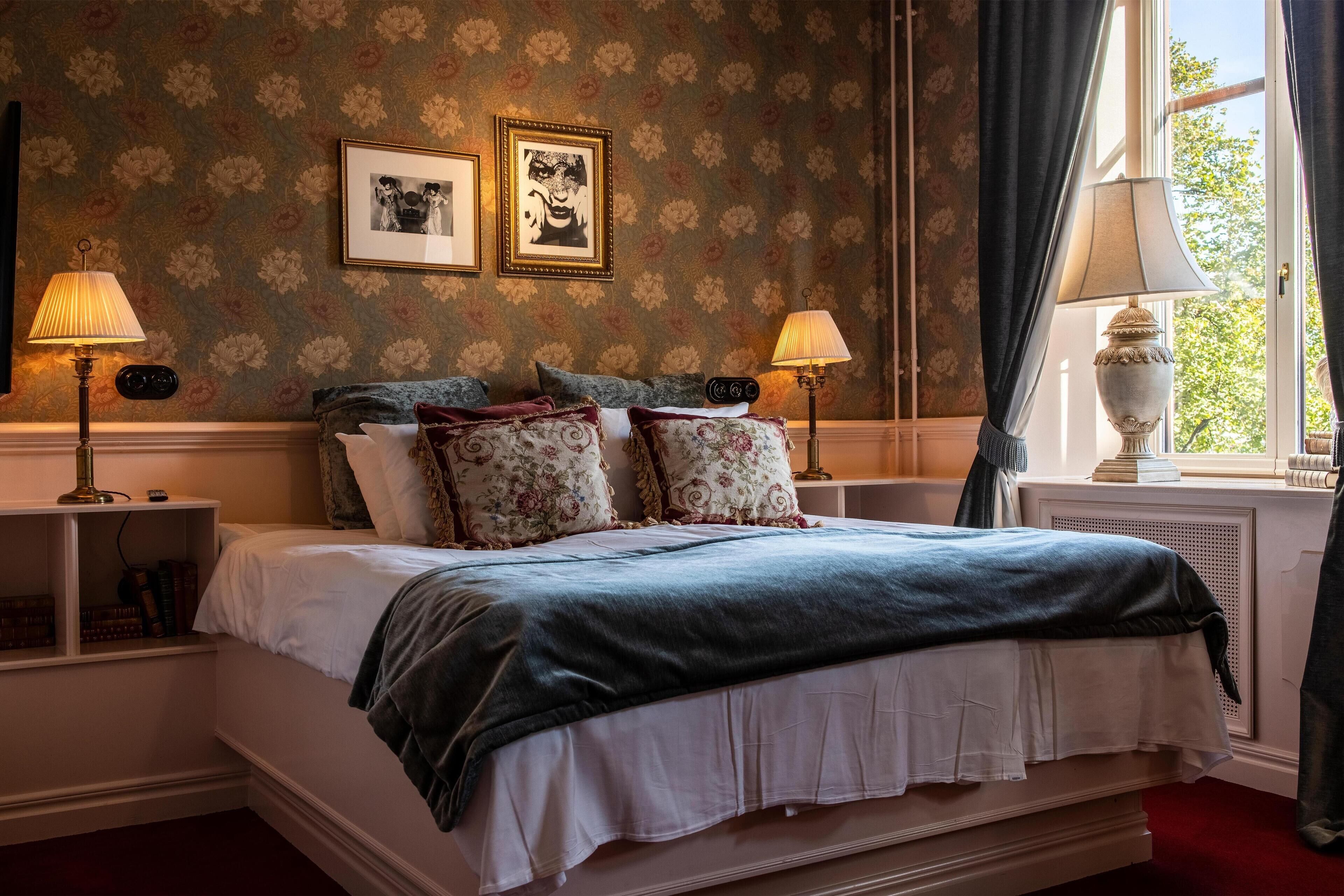 A classic bedroom with a large bed, floral wallpaper, two framed pictures, and two bedside lamps.