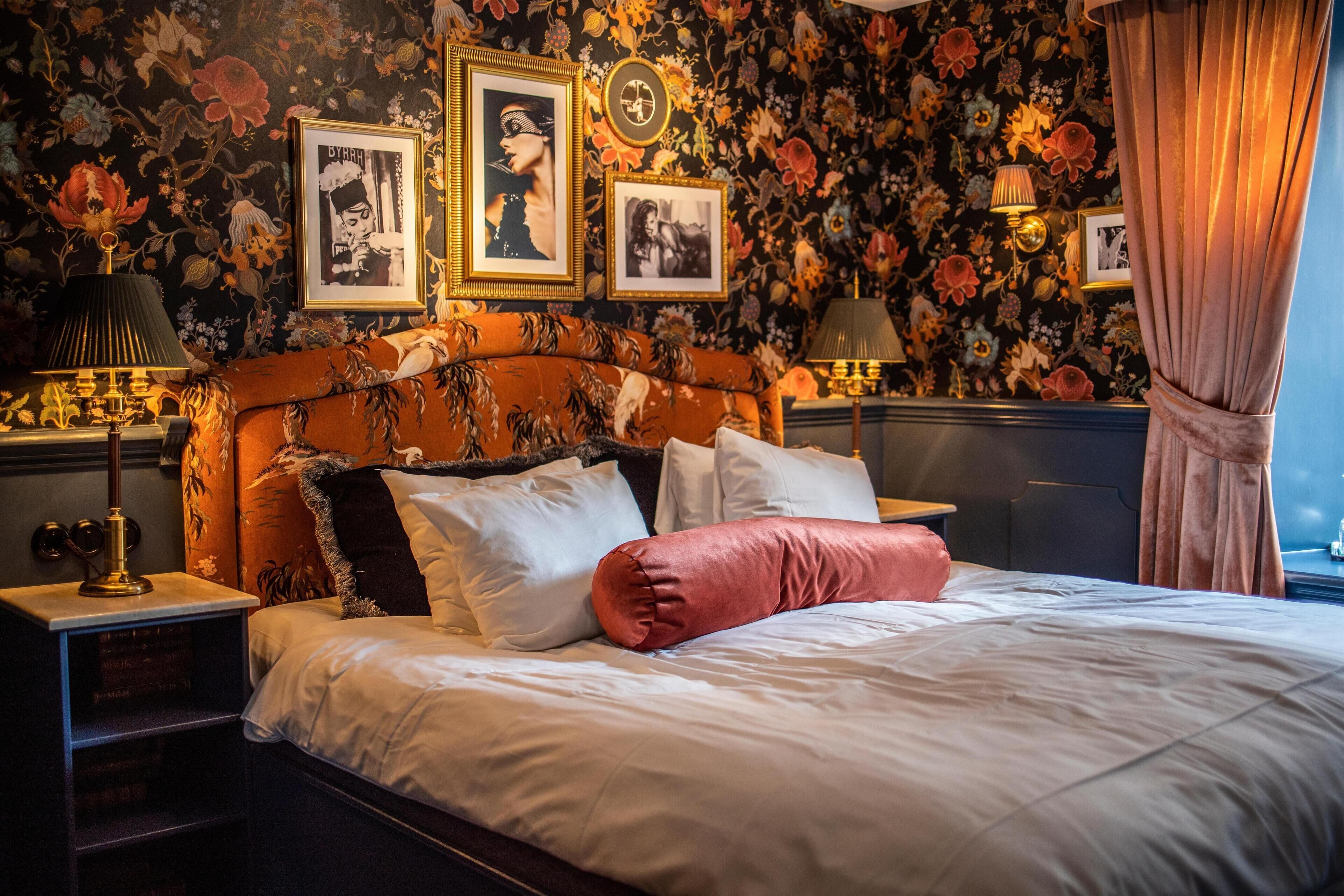 A bed with white bedding and a pink pillow, flanked by two nightstands with lamps. The room features floral wallpaper and framed pictures on the wall.