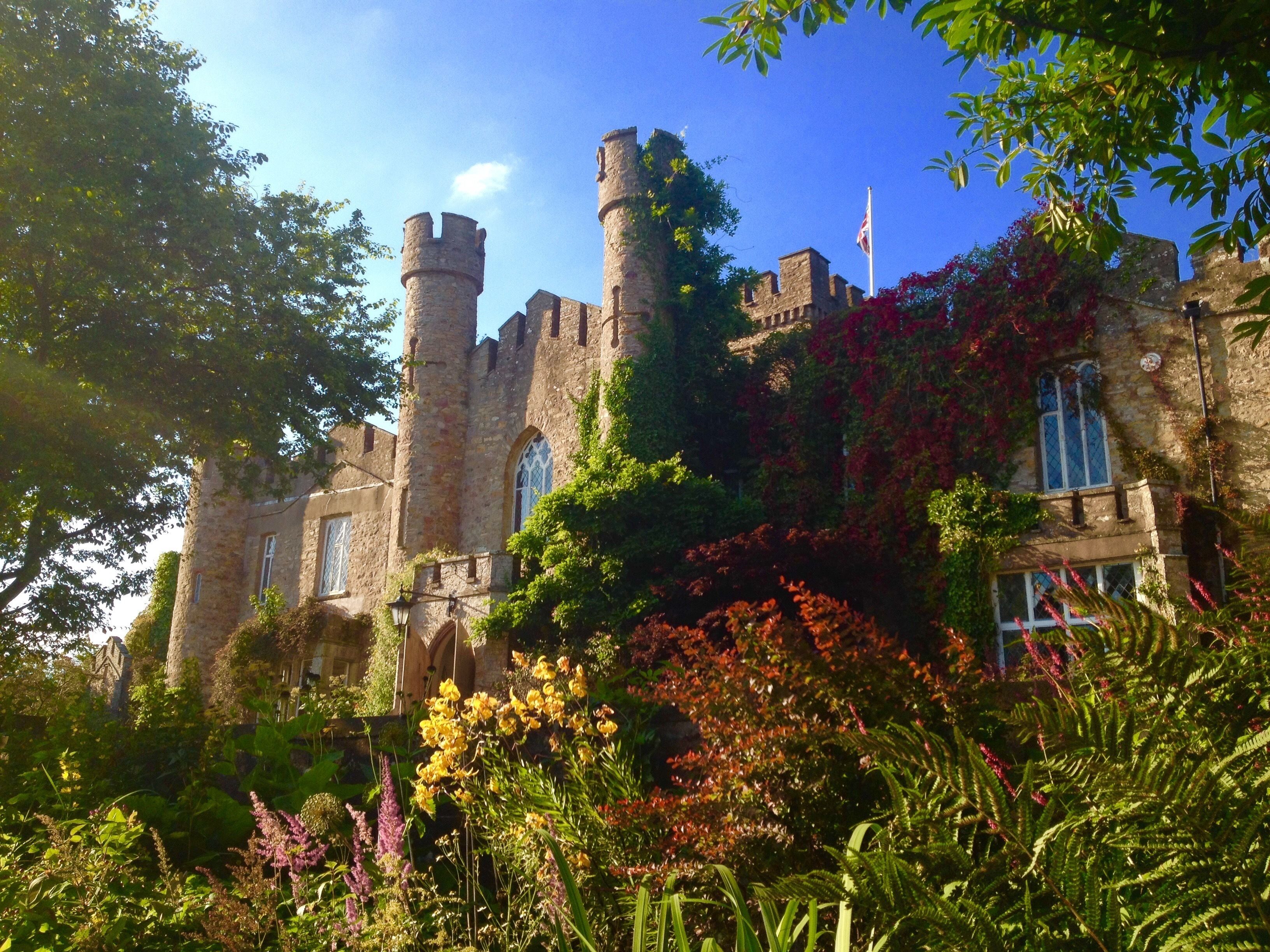 A historic stone castle with multiple towers, surrounded by lush greenery and flowering plants.
