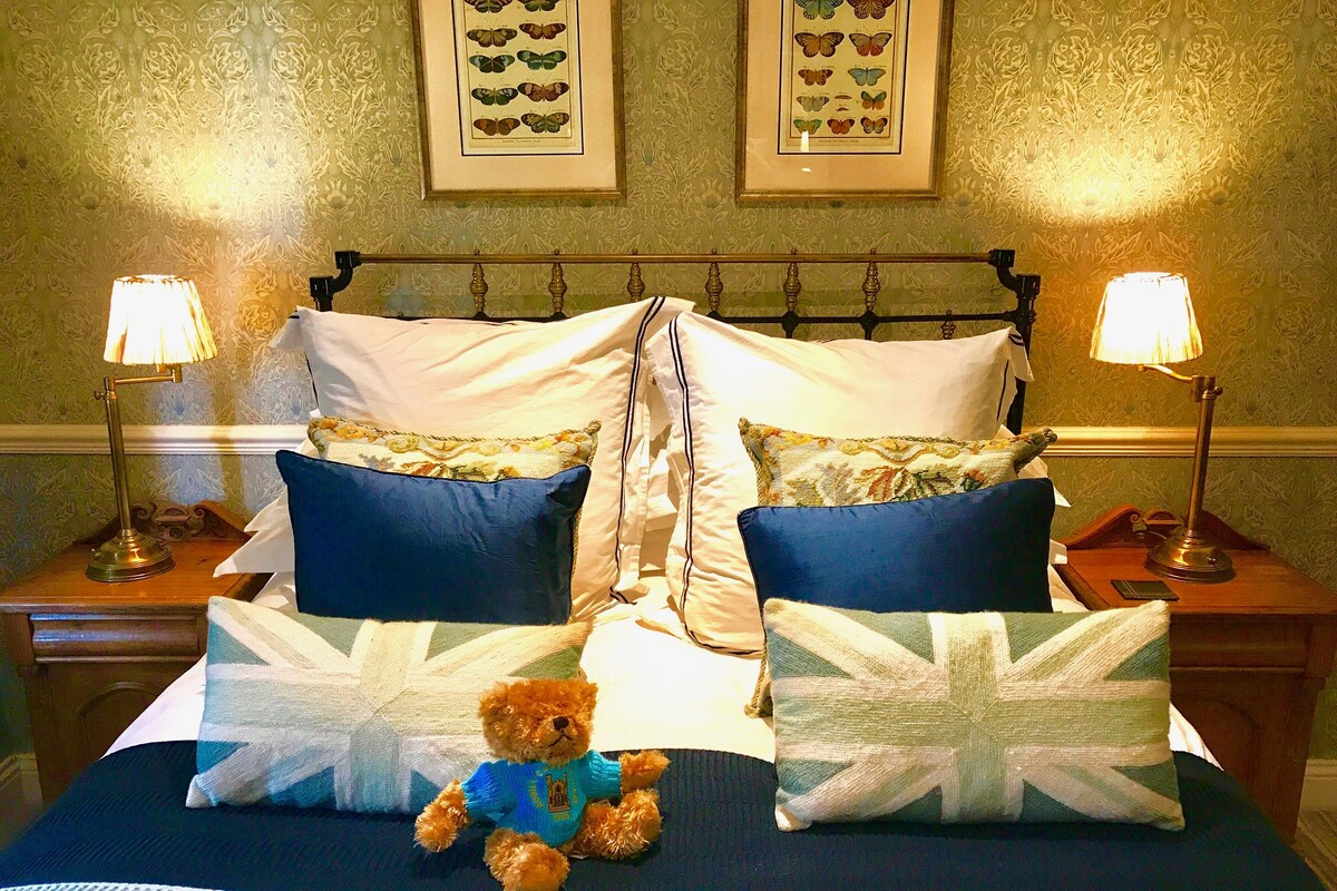 Una cama bien hecha con un osito de peluche, dos lámparas y cuadros de mariposas en la pared.
