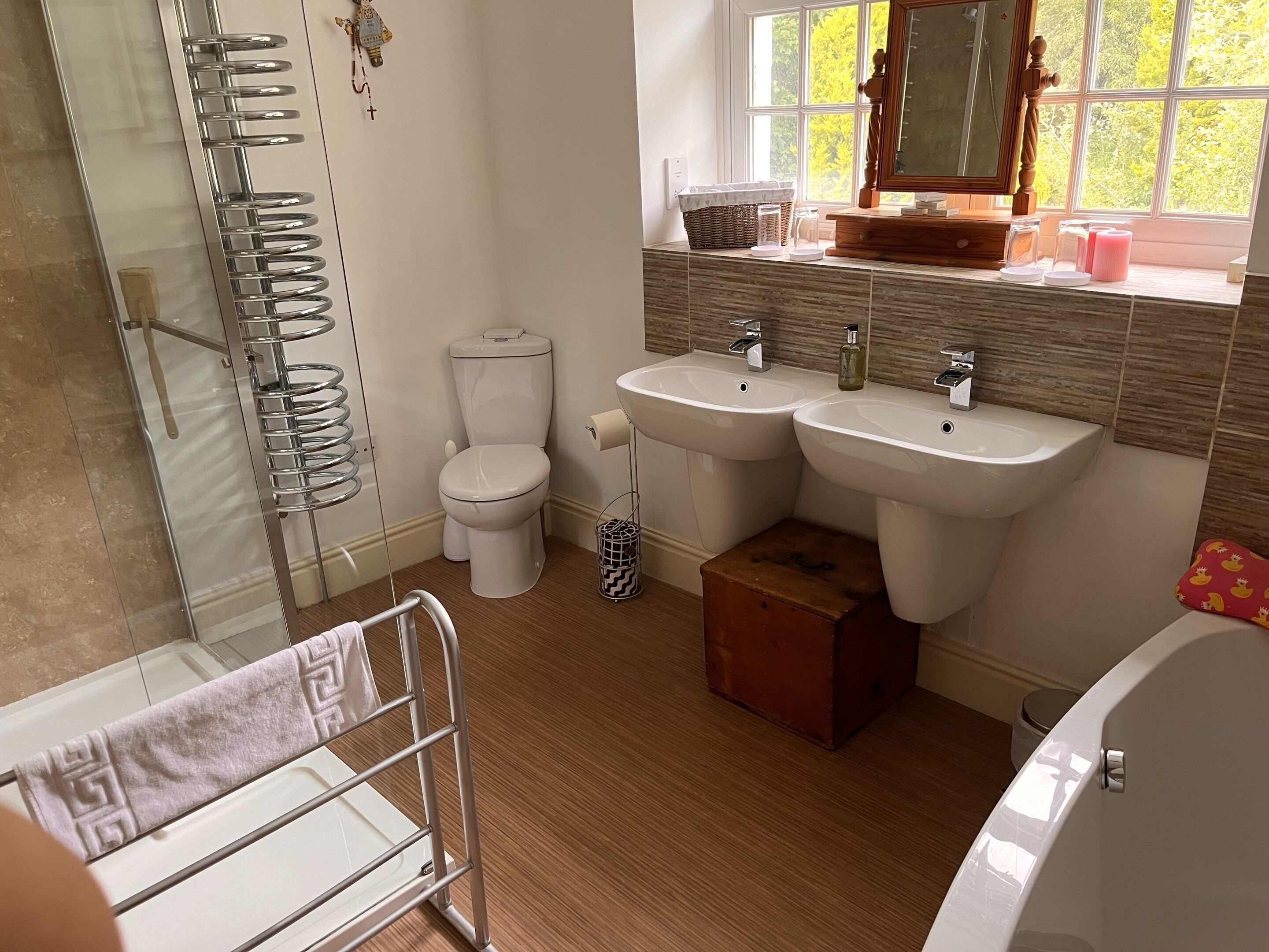 A bathroom with a glass shower enclosure, a toilet, two sinks, a wooden vanity, and a window with a view of trees.