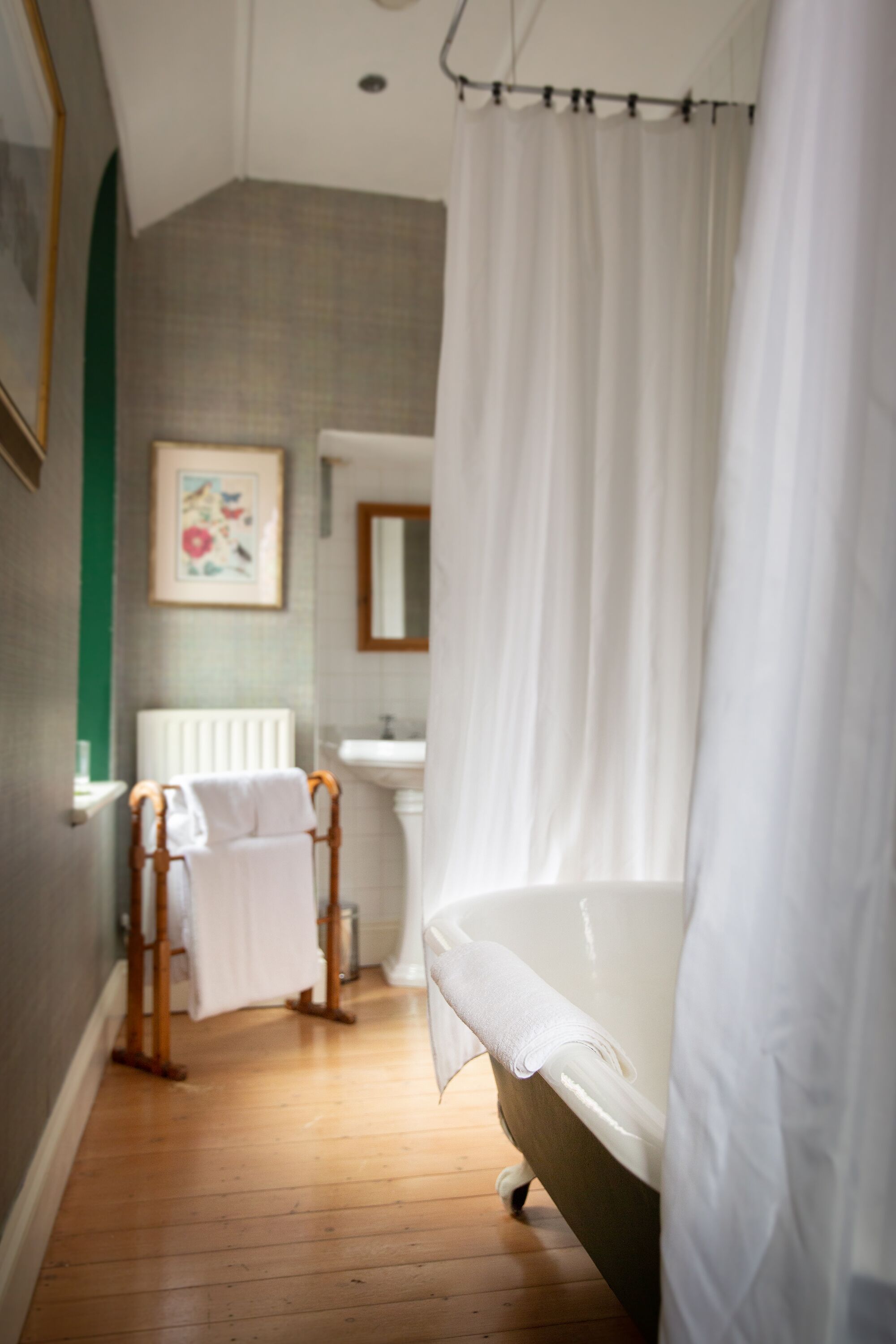 A bedroom with a wooden floor, a bed with a white sheet, a white curtain, a radiator, a sink, and a framed picture on the wall.