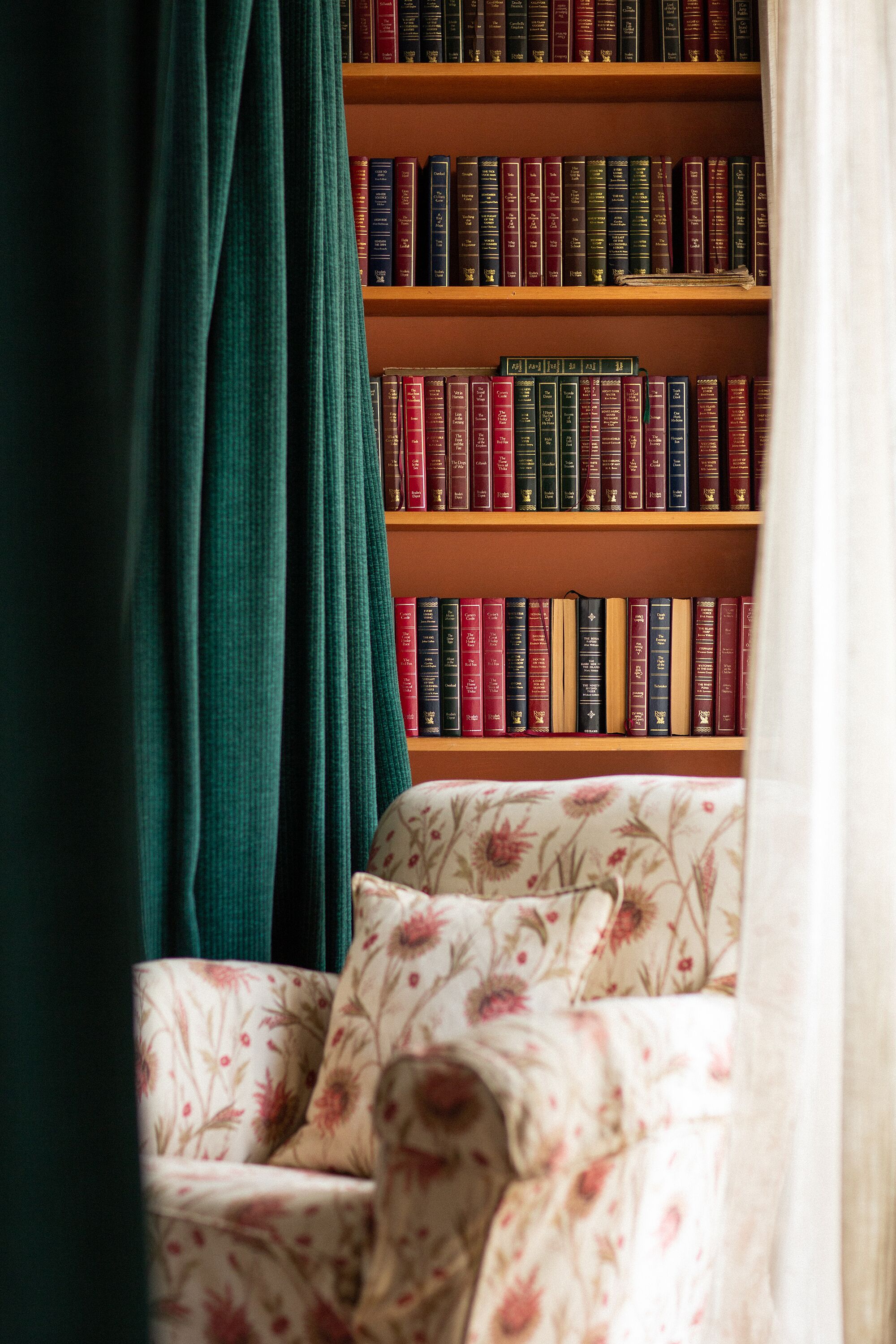 Una libreria piena di libri, una poltrona con motivo floreale e una tenda verde che copre parzialmente la vista.